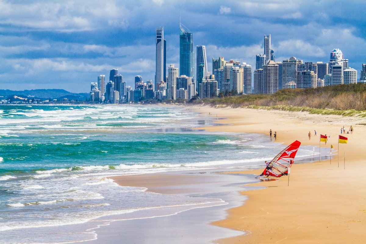 View of Surfers Paradise on Queensland’s Gold Coast, with turquoise waves rolling onto a long sandy beach, a red windsurf sail near the shoreline, and the high-rise skyline under a cloudy sky.